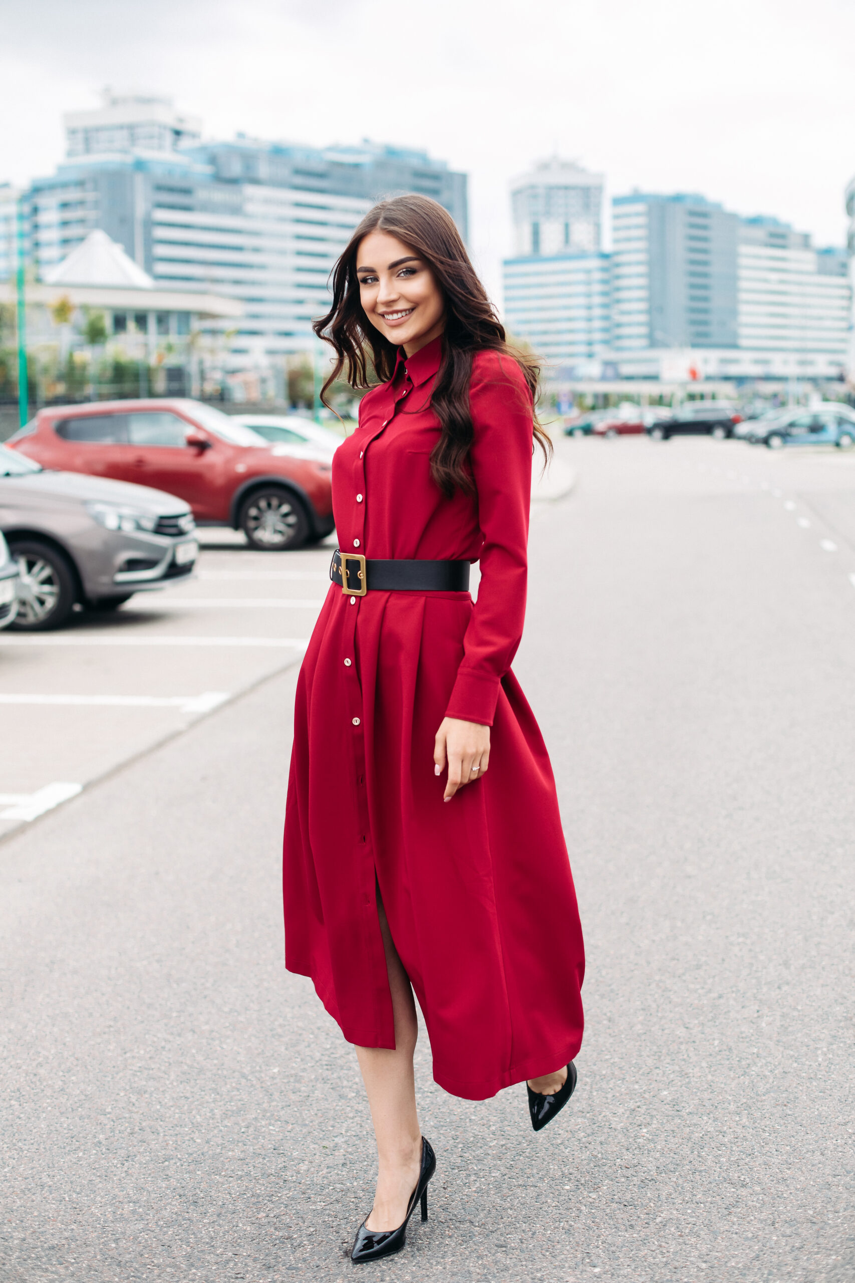 Happy pretty young lady in red dress looking at camera while walking along the street in modern city. Lifestyle concept